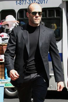 a man in a black suit and sunglasses walking down the street next to a bus