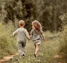 two young children walking down a dirt path in the middle of tall grass and trees