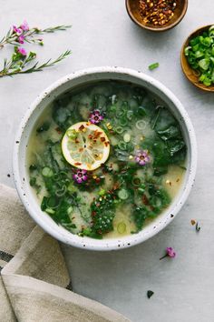 a white bowl filled with soup next to two bowls of greens and lemon wedges