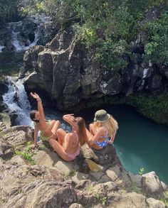 two women in bikinis are sitting on rocks near a waterfall and looking at the water