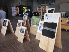 three wooden easels with paintings on them and people looking at them in the background