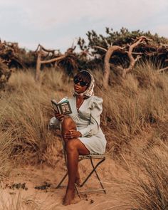 a woman sitting in a chair on the beach reading a book while wearing a headscarf