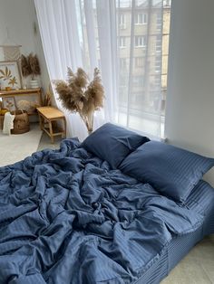 an unmade bed with blue sheets and pillows in front of a large window overlooking the city