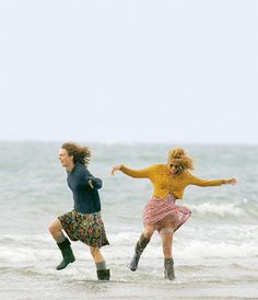 two women are running in the water at the beach, one is wearing rain boots