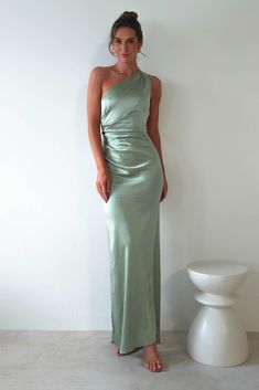 a woman standing in front of a white wall wearing a green one - shoulder dress