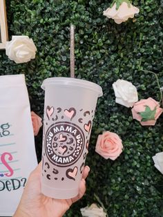 a person holding up a cup with the word mama on it and flowers in the background