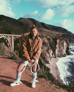 a woman standing on the side of a cliff next to the ocean with a bridge in the background