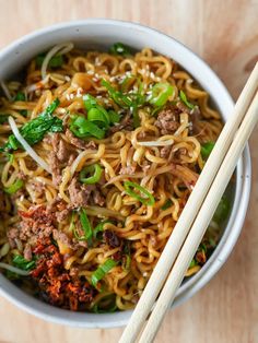 a bowl filled with noodles, meat and vegetables next to chopsticks on a wooden table