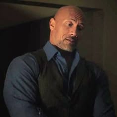 a bald man in a blue shirt and black vest is looking at the camera with his hands on his hips