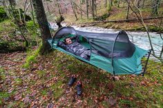 a man laying in a sleeping bag next to a river with trees and leaves on the ground