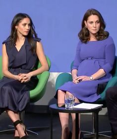 two women sitting next to each other on chairs in front of a blue wall and one woman wearing a purple dress