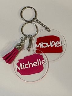 two acrylic key chains with tassels attached to them, one is red and the other is pink