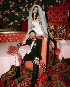 a bride and groom sitting on a red chair in front of a table with pink flowers