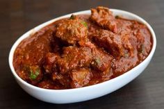 a white bowl filled with meat and sauce on top of a wooden table next to a fork