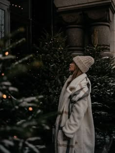 a woman standing in front of a christmas tree wearing a white coat and plaid hat