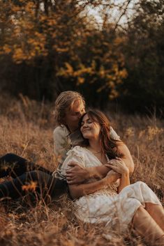 a man and woman sitting on the ground in tall brown grass with trees behind them