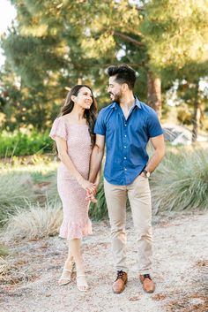 an engaged couple holding hands and smiling at each other while standing in the sand near some trees