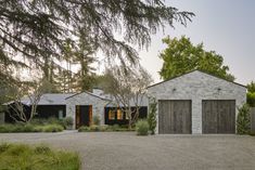 a stone house with two garages and trees in the front yard, surrounded by greenery