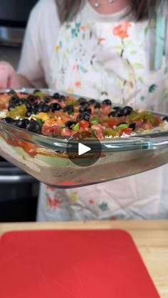 a woman is holding up a casserole dish with black olives and peppers