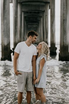 a man and woman standing in the water under a pier with their arms around each other