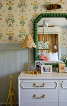 a dresser with a mirror, lamp and pictures on top of it in front of a wallpapered bedroom