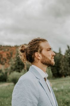 a man with a messy ponytail standing in a field looking off into the distance while wearing a blue jacket