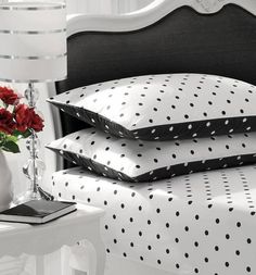 three pillows on a white bed with black and white polka dot print, one has red roses in a vase next to it