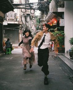 a man and woman running down the street in an alleyway with buildings on either side