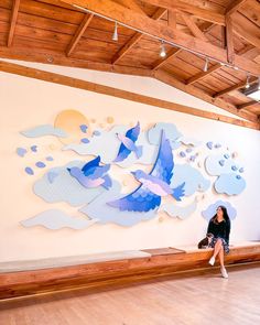 a woman sitting on a bench in front of a wall with birds and clouds painted on it