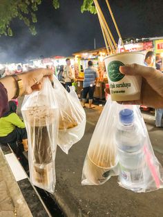 a person holding two plastic bags with food in them and people standing around at an outdoor market