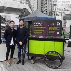 two people standing in front of a cart with information on it and an umbrella behind them