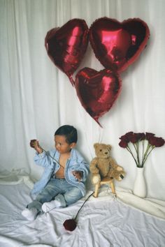 a little boy sitting on top of a bed next to two red heart shaped balloons