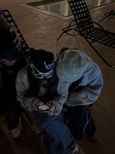 two people sitting on a bench looking at their cell phones in front of an empty swimming pool