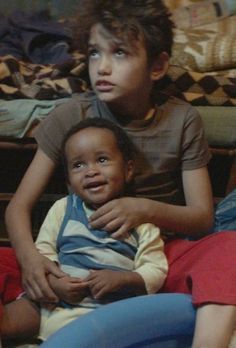 two young children sitting on top of a bed with their arms around each other's shoulders