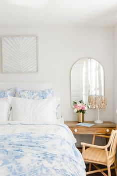 a white bed with blue and white comforter next to a wooden chair in front of a mirror