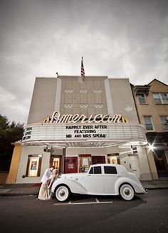 two people standing in front of a movie theater with an old car parked on the street