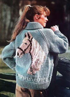 a woman standing next to a wooden fence wearing a sweater with a horse on it