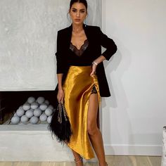 a woman standing in front of a fireplace wearing a gold skirt and black blazer