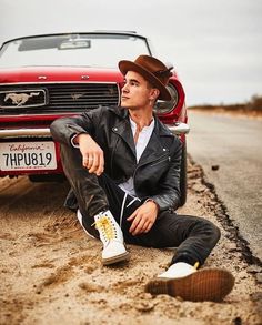 a man sitting on the ground next to a red car and wearing a brown hat