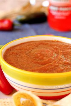 a yellow and white bowl filled with red sauce next to crackers, tomatoes and peppers