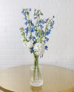 blue and white flowers in a clear vase on a gold table with brick wall behind it