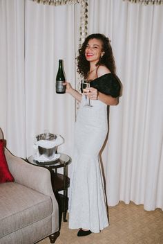 a woman in a white dress holding a wine bottle and glass while standing next to a couch