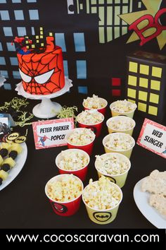 a table topped with cups filled with popcorn and spiderman cake next to other desserts