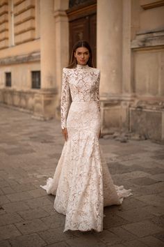 a woman standing in front of a building wearing a wedding dress with long sleeves and high neck