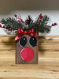 a wooden block with a reindeer face and red nose on it, decorated with evergreen branches