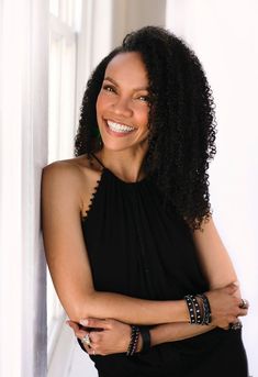 a smiling woman in a black dress leaning against a wall with her hands on her hips