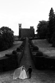 a bride and groom walking in front of an old castle with hedges on either side