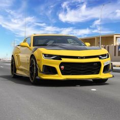 a yellow chevrolet camaro is driving down the road in front of a building with blue sky and clouds