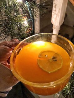 a person holding up a glass with a lemon slice on it in front of a christmas tree