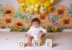 a baby sitting on the floor with blocks spelling out one, surrounded by balloons and flowers